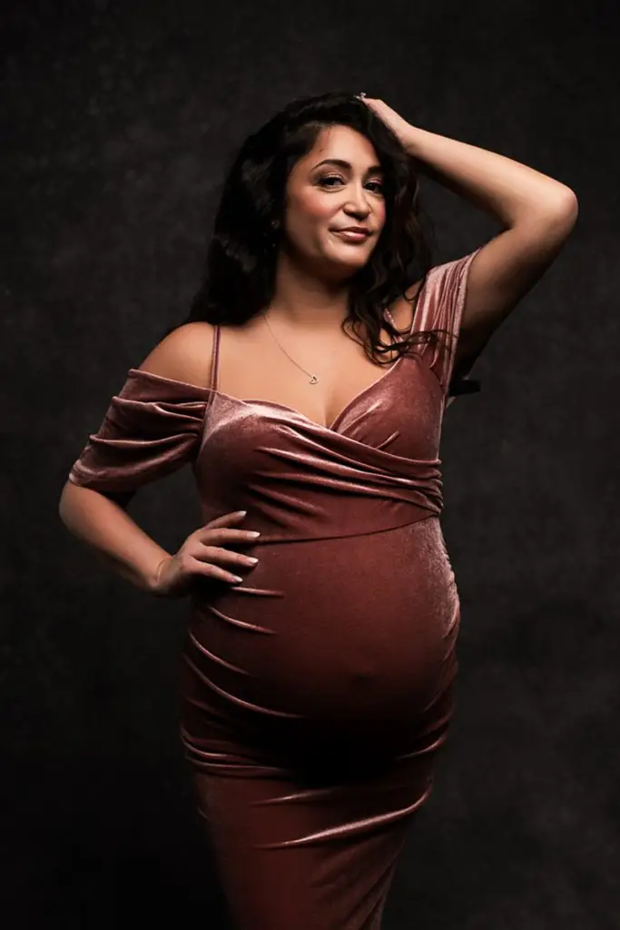 Pregnant woman in a mauve velvet dress poses confidently, one hand on her hip, other touching her wavy hair, against a textured dark background.