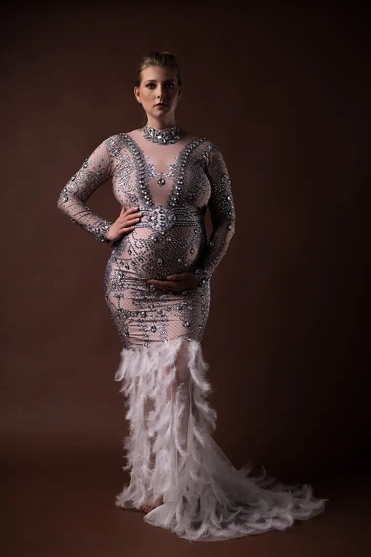 Pregnant woman in an ornate, jeweled, sheer gown with feather details stands confidently against a dark brown background, exuding elegance and grace.