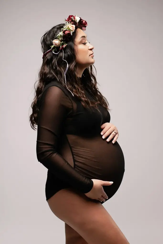 Pregnant woman in a black sheer dress, side view, cradles her belly and wears a floral crown. Her expression is serene, set against a gray backdrop.