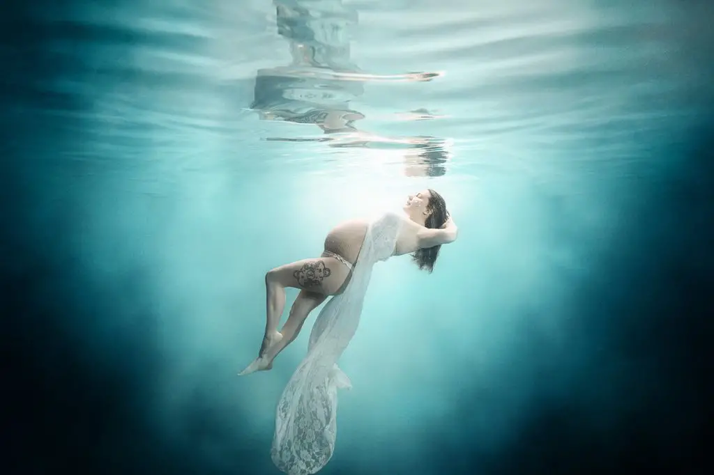 Pregnant woman floats underwater, wrapped in a sheer white fabric, with serene expression. Soft blue tones create a tranquil, ethereal atmosphere.