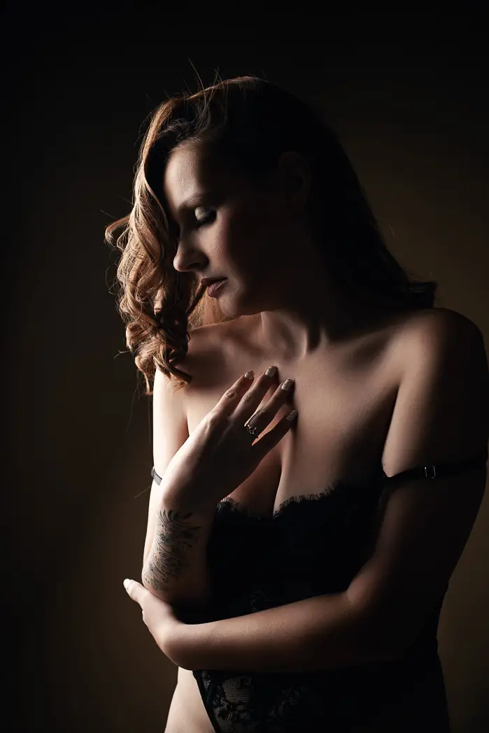 A woman in black lace lingerie is softly lit from the side, creating a dramatic shadow. Her hand is on her chest, and her expression is serene and introspective.