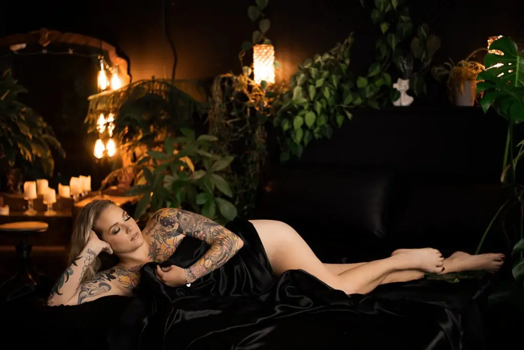 Studio vs Outdoor Boudoir in Syracuse NY. Girl lying on a bed draped in a black silk blanket with candles and plants in the background.
