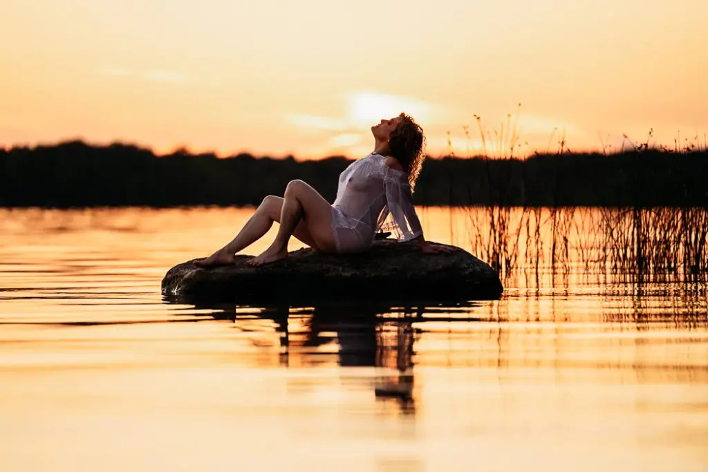 Girl posing in a light shear lingerie top on a rock rising out of the lake, siren-like, with the sunset in the background. Outdoor Boudoir Syracuse NY