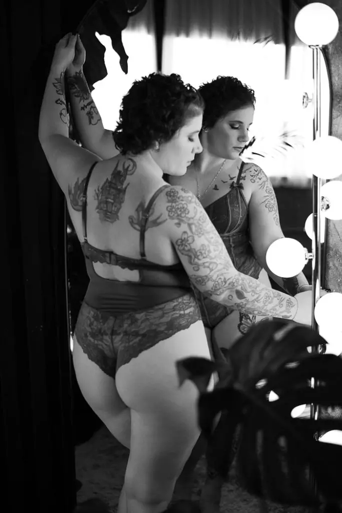 Studio vs Outdoor Boudoir in Syracuse NY. Black and white photo of a girl posing in front of a mirror in lingerie while looking down and to the right of the mirror.